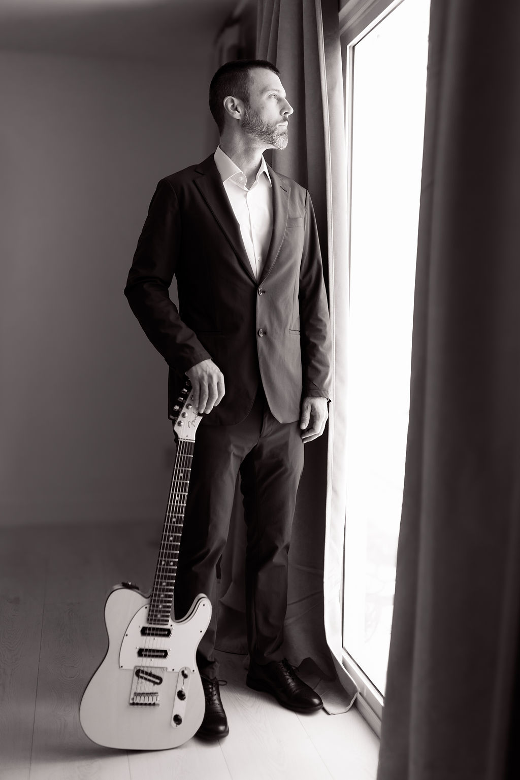 Coyah Gamble in a suit holding his guitar, looking out a window